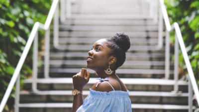Woman standing with profile view and fisted hand brought to chin at bottom of outdoor stairs